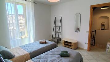 A bedroom with two beds, a ceiling fan, a window with curtains, a wooden bench, and a mirror.