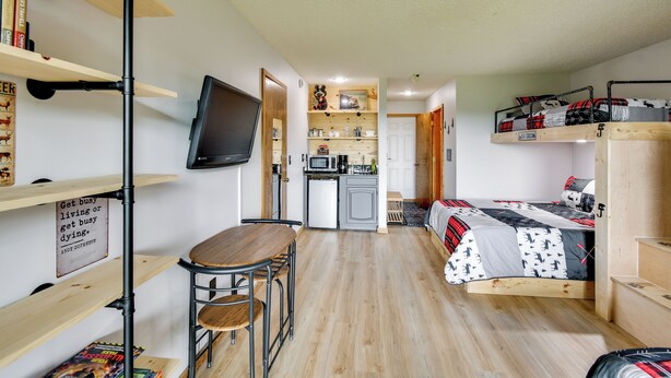 Quarto compacto com cama, pequena cozinha, área de refeições e televisão na parede.
