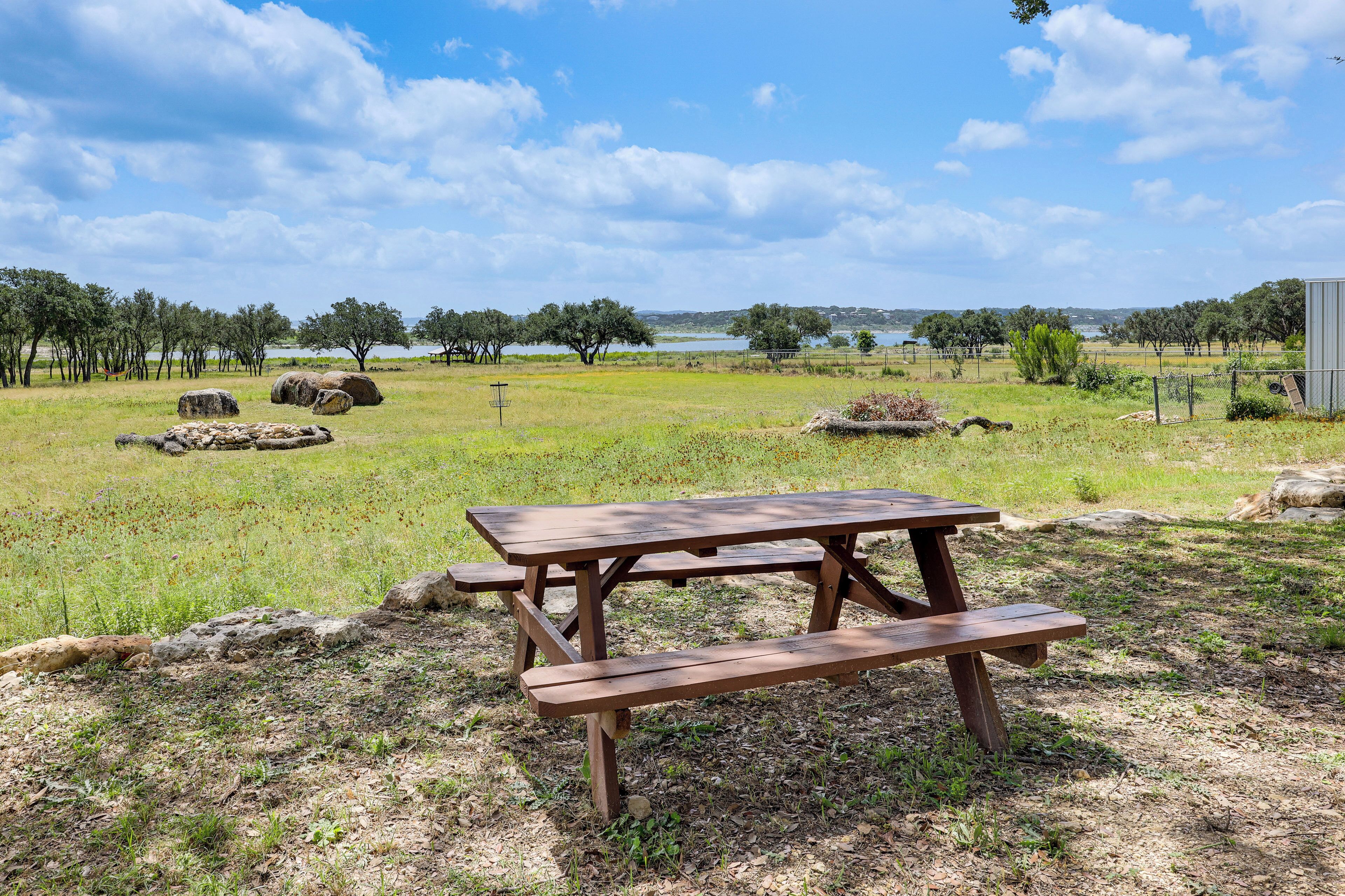 Une table de pique-nique dans un champ herbeux, avec des arbres et une étendue d’eau en arrière-plan.