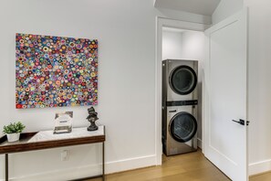 Ein Waschraum mit einer großen, an der Wand montierten Waschmaschine und einem Trockner, einem kleinen Tisch mit einer Pflanze und einem farbenfrohen, abstrakten Gemälde darüber.