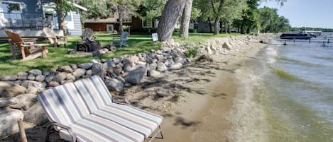 A lakeside property with a sandy beach, a row of Adirondack chairs, and a row of striped lounge chairs.