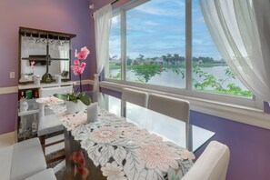 Un comedor con una mesa dispuesta para dos personas, una ventana con vistas al agua y vegetación, y un espejo con copas colgantes.