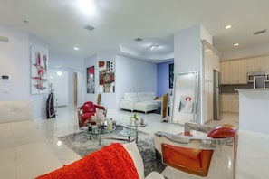 Una sala de estar moderna con un sofá blanco, sillas rojas, una mesa de centro de vidrio y una zona de cocina con armarios y electrodomésticos.
