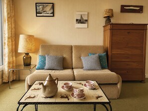 A cozy living room with a sofa, a side table with a lamp, a dresser, and a table with teapots and cups. - Les Guillemots by Interhome (Blonville sur mer)