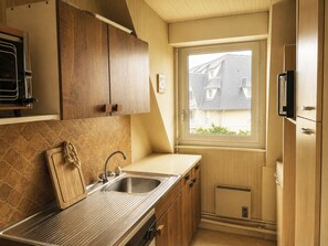 A kitchen with wooden cabinets, a stainless steel sink, a window with a view of buildings, and a built-in oven. - Les Guillemots by Interhome (Blonville sur mer)