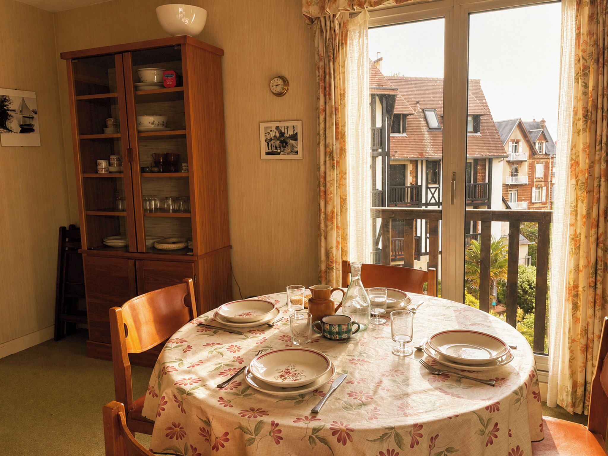 Ein Essbereich mit einem runden Tisch für zwei Personen, einem Holzschrank mit Geschirr und einem Balkon mit Blick auf Häuser.