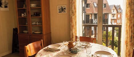 Un coin repas avec une table ronde dressée pour deux personnes, un buffet en bois contenant de la vaisselle et un balcon offrant une vue sur les maisons.