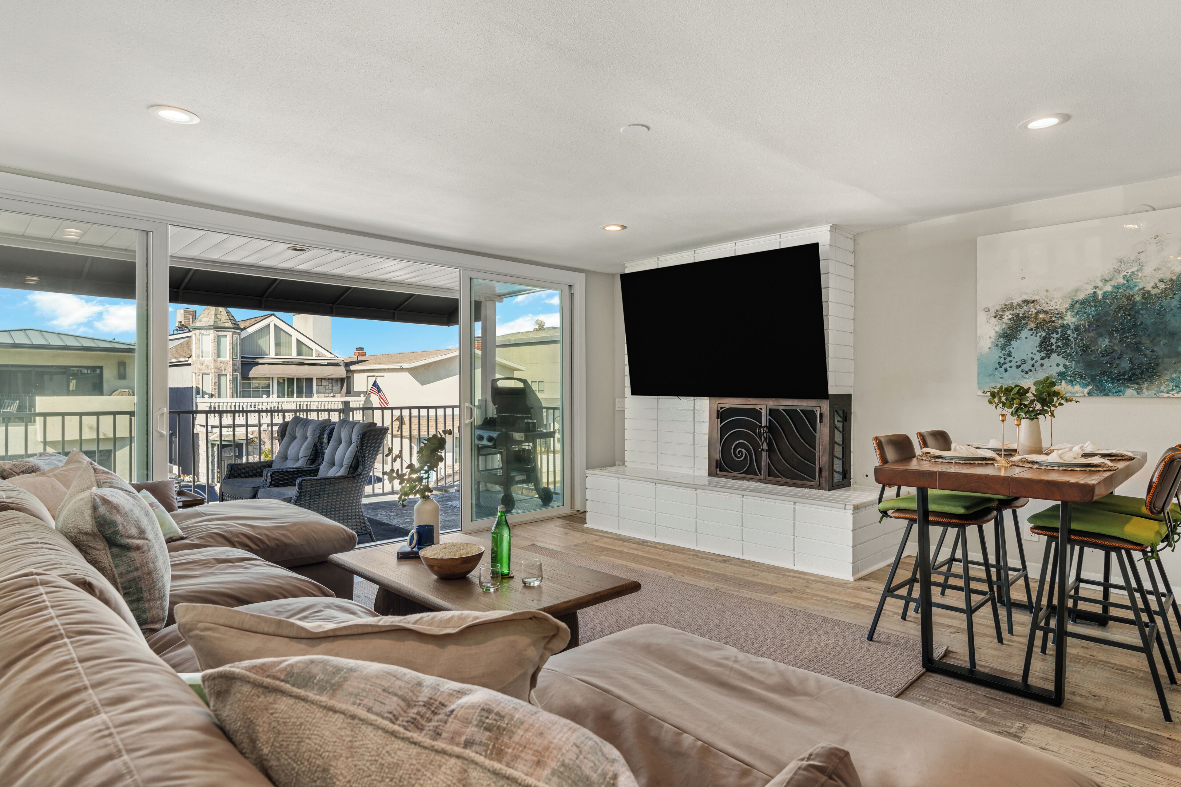 Uma sala de estar moderna com um sofá grande, uma mesa de jantar com cadeiras, uma lareira e uma janela ampla com vista para uma área residencial.