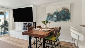 A dining area with a wooden table set for four, surrounded by chairs with green and brown upholstery. A large abstract painting adorns the wall above the table, and a flat-screen TV is mounted on the wall. - Entire Duplex + Steps to Beach | Newport Beach | Harborlight & Seabright Sky (Newport Beach)