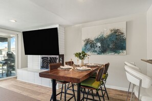 Ein Essbereich mit einem Holztisch, gedeckt für vier Personen, umgeben von Stühlen mit grün-brauner Polsterung. Über dem Tisch hängt ein großes abstraktes Gemälde, und an der Wand ist ein Flachbildfernseher angebracht.