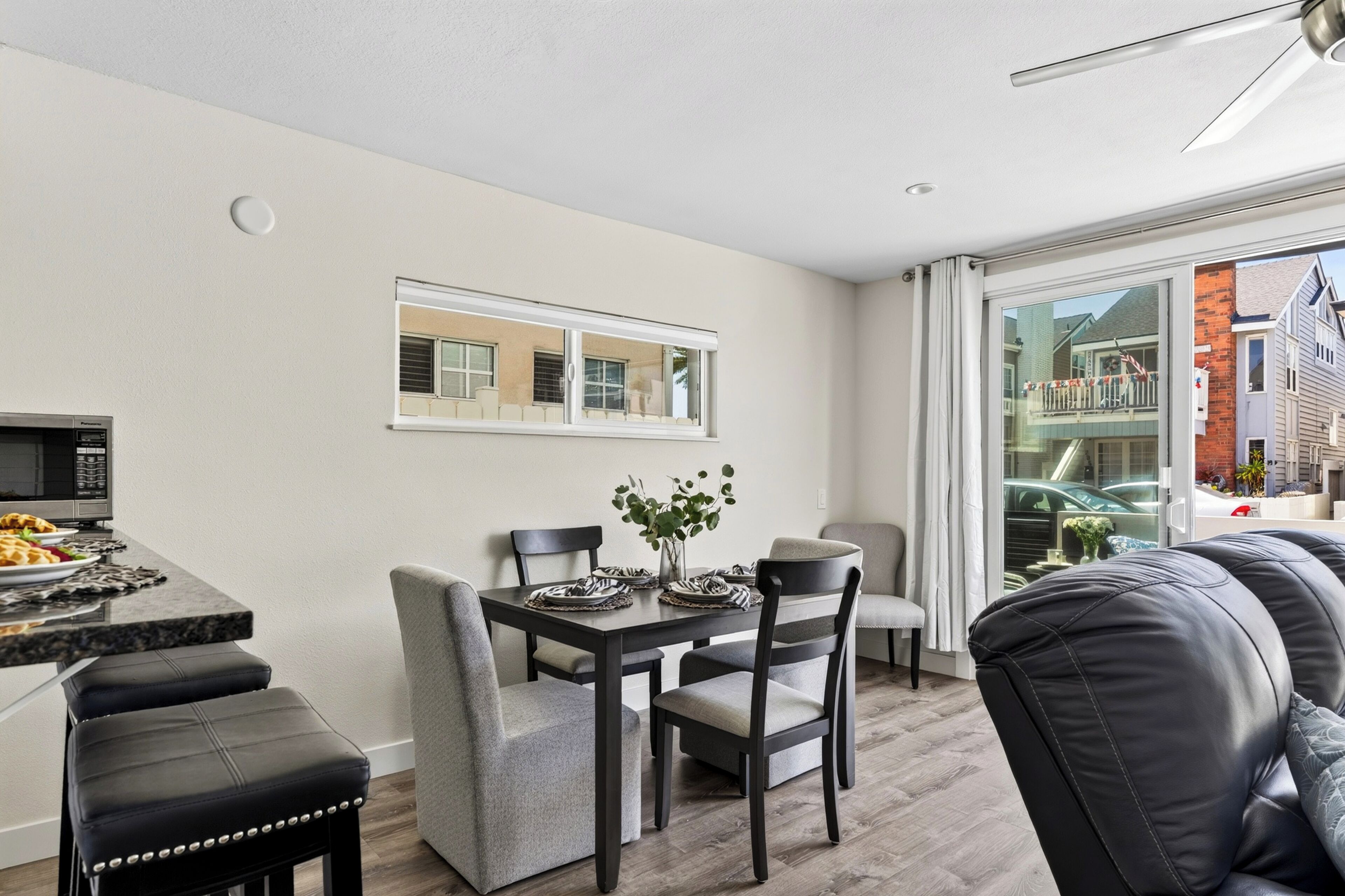 A modern dining area with a table set for two, a black leather bench, and a view of a residential building through a glass door.