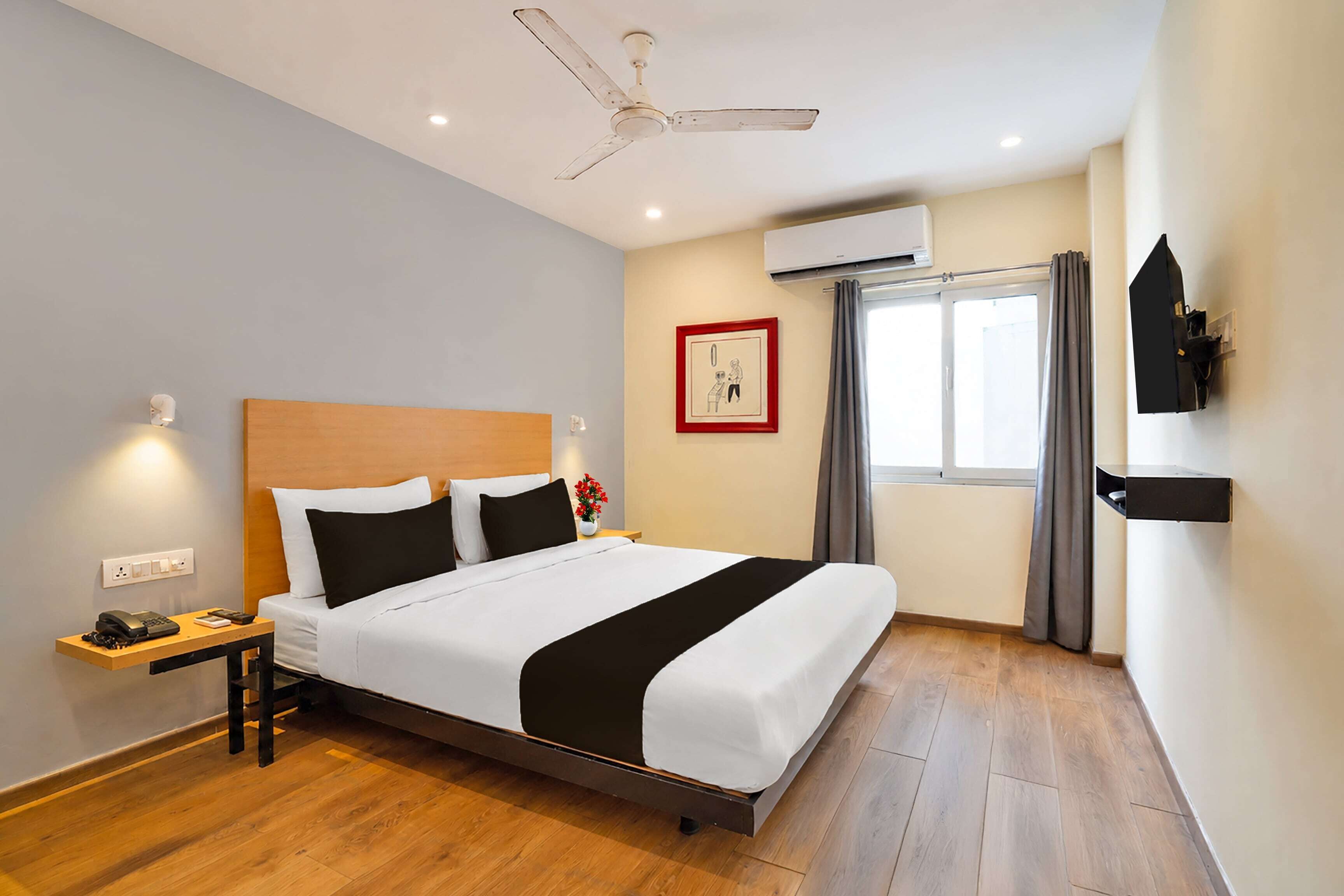 A hotel room with a bed, bedside table, nightstand, wall-mounted TV, ceiling fan, window with curtains, and an air conditioning unit.