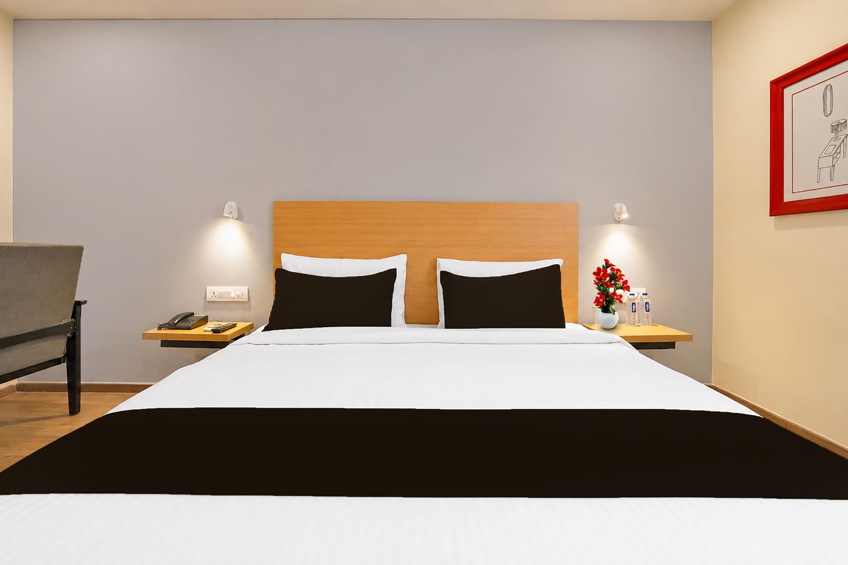 A hotel room with a large bed, two bedside tables, a chair, a nightstand with a phone, a vase with flowers, and a framed picture on the wall.