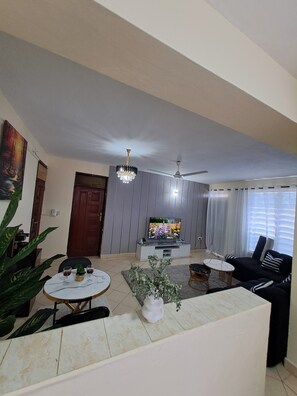 Ein Wohnzimmer mit Fernseher, Deckenventilator, einer Kandelaberleuchte und einem Essbereich mit Tisch und Stühlen.