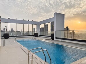 A rooftop pool with a city view, surrounded by glass railings and large planters. - FIRST CLASS | 1BR | Modern Haven | Tranquil City (JVC)