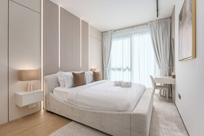 A modern hotel room with a bed, bedside table, lamp, and a desk with a chair. - FIRST CLASS | 1BR | Modern Haven | Tranquil City (JVC)