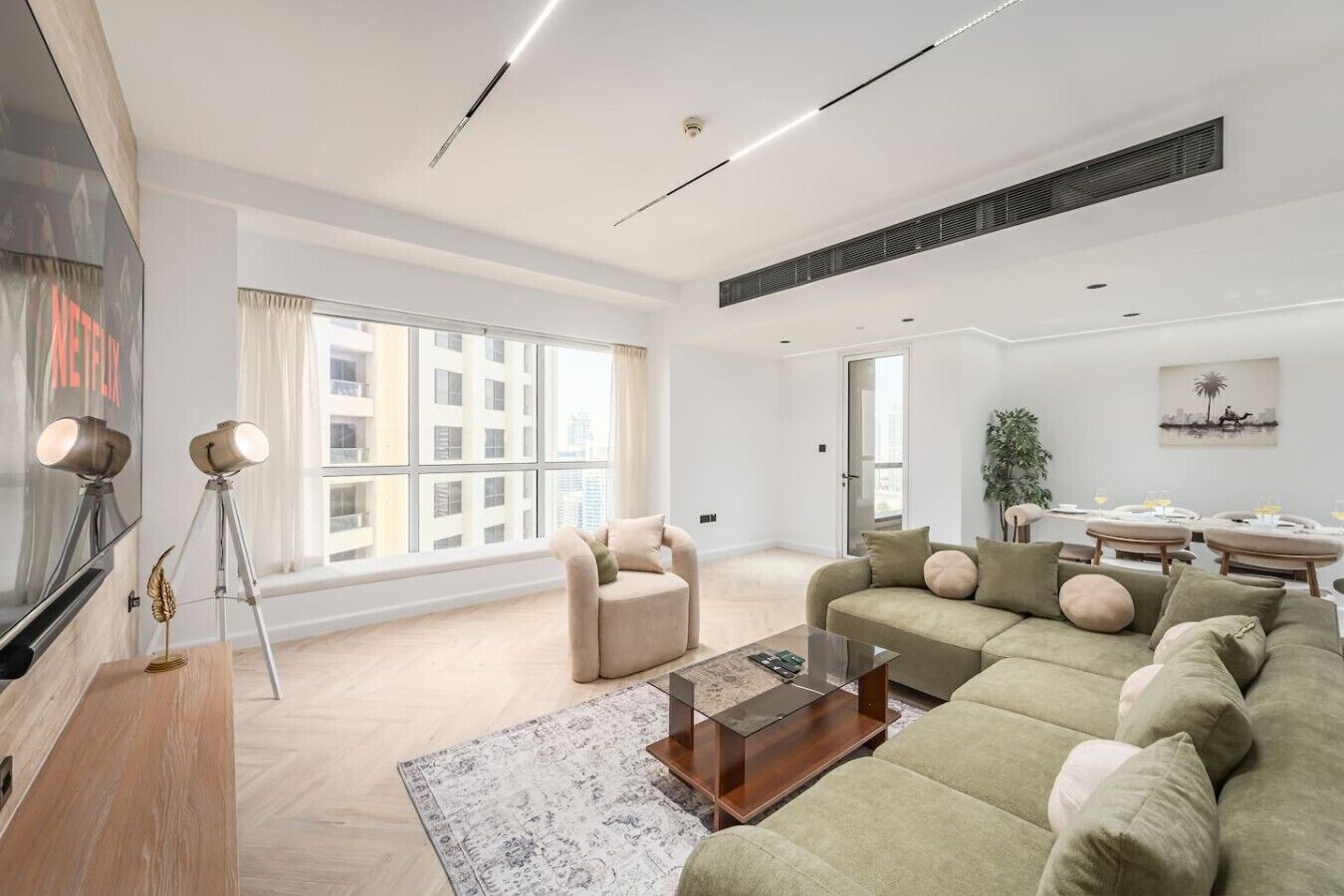 A modern living room with a sofa, armchair, and coffee table, large windows, and a dining area with a plant.