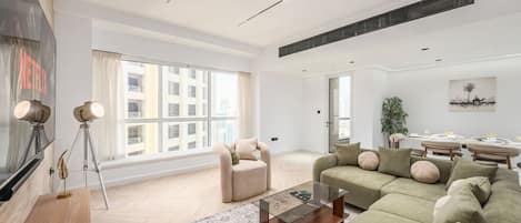 A modern living room with a sofa, armchair, and coffee table, large windows, and a dining area with a plant.