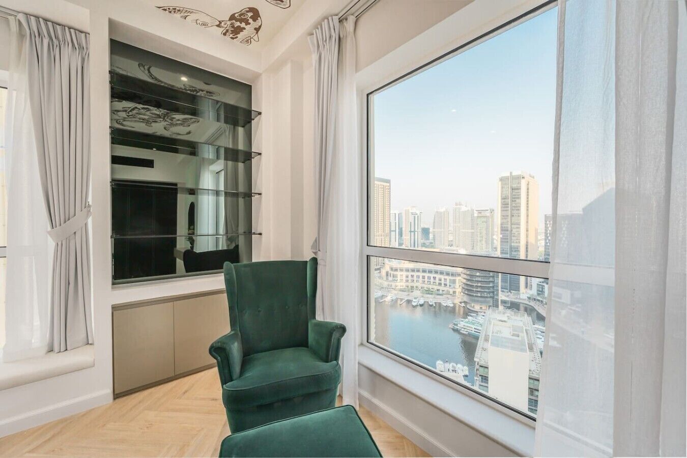 A room with a large window, a green chair, and a glass shelving unit with decorative items.