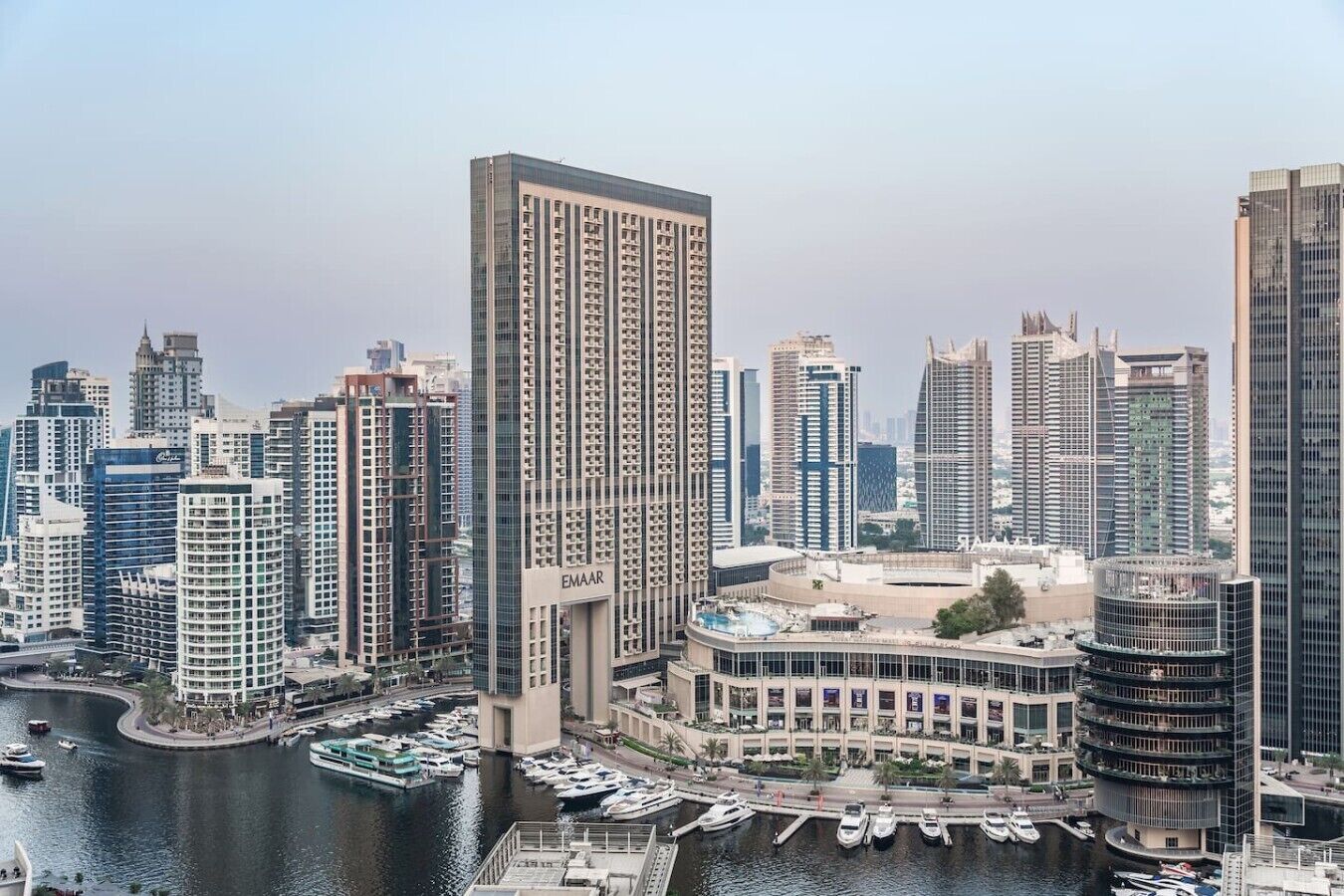 Un edificio de gran altura con un llamativo letrero 'EMAAR', rodeado de otros edificios altos, un puerto deportivo con barcos y un paseo marítimo.