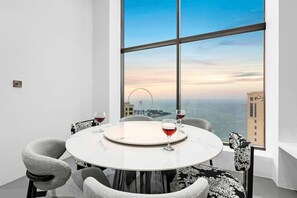 A round table with a white marble top, two chairs with grey upholstery, and two wine glasses with red wine. A large window reveals a cityscape with a Ferris wheel and buildings. - FIRST CLASS |3BR| Luxury Duplex w/ Ain Dubai View (JBR)