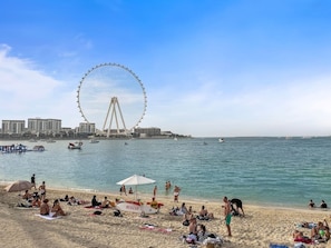 A beach with people sunbathing, a Ferris wheel in the background, and boats on the water. - FIRST CLASS |3BR| Luxury Duplex w/ Ain Dubai View (JBR)
