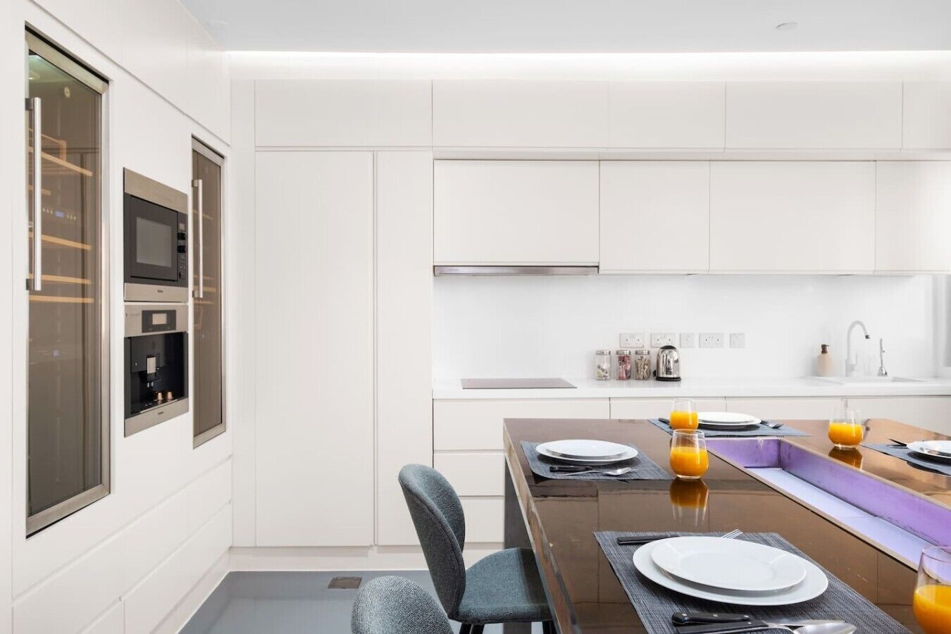 Une cuisine moderne dotée d’appareils électroménagers intégrés, un coin repas avec une table dressée pour deux personnes et une grande fenêtre.