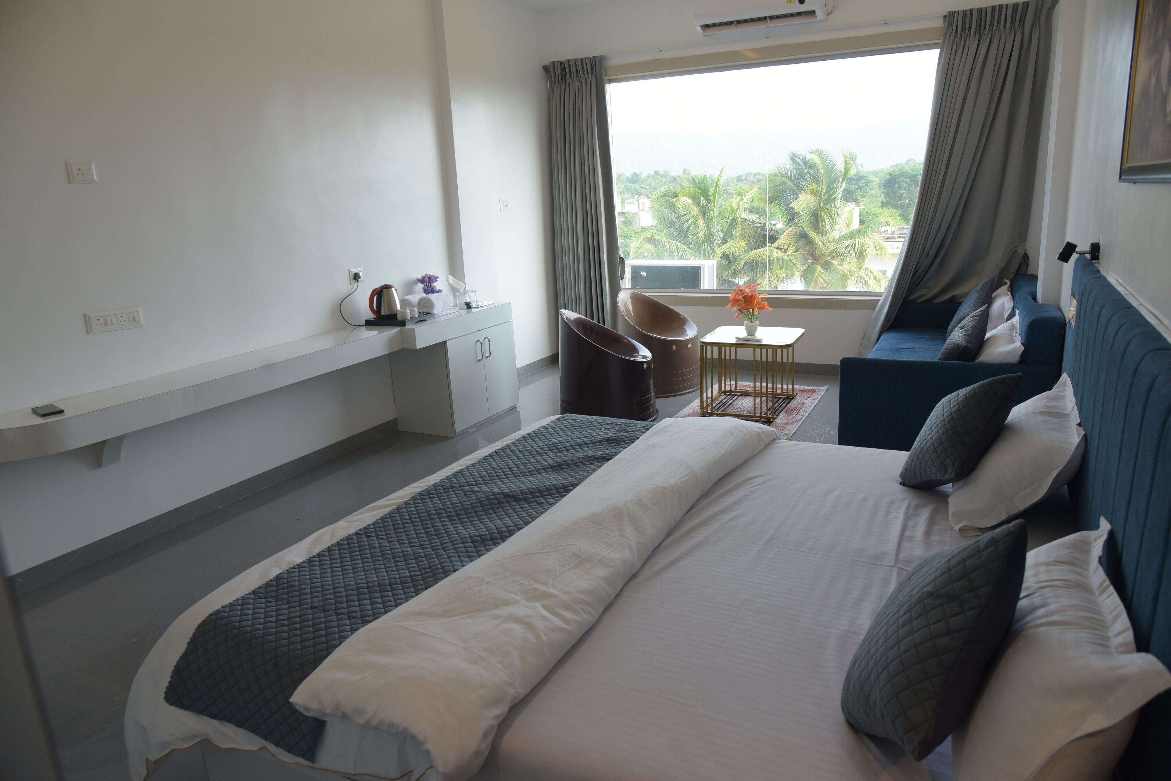 A hotel room with a bed, a sofa, a small table with a vase, a chair, and a window with curtains.