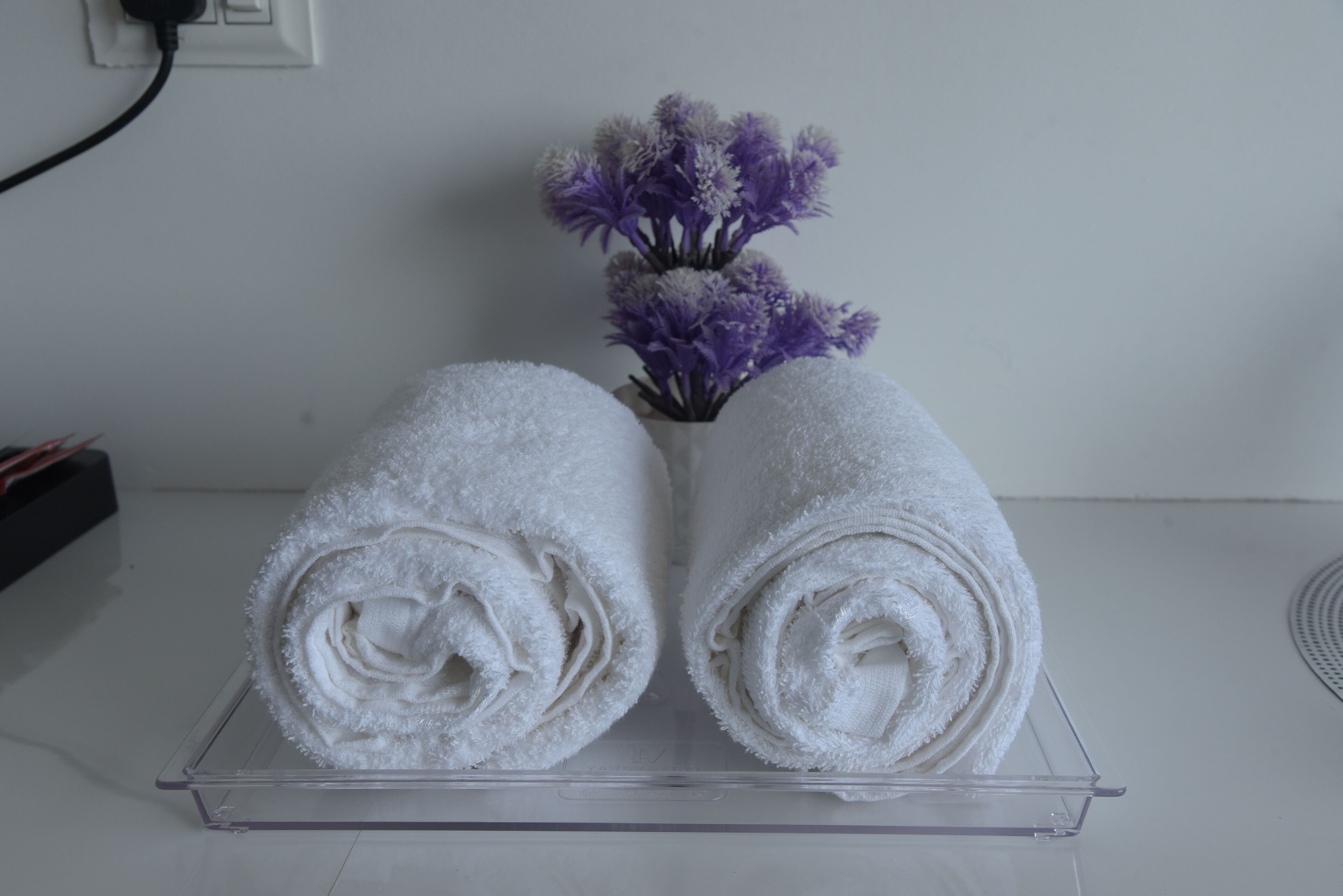 Deux serviettes blanches roulées, posées sur un plateau transparent, avec un vase de fleurs violettes en arrière-plan.