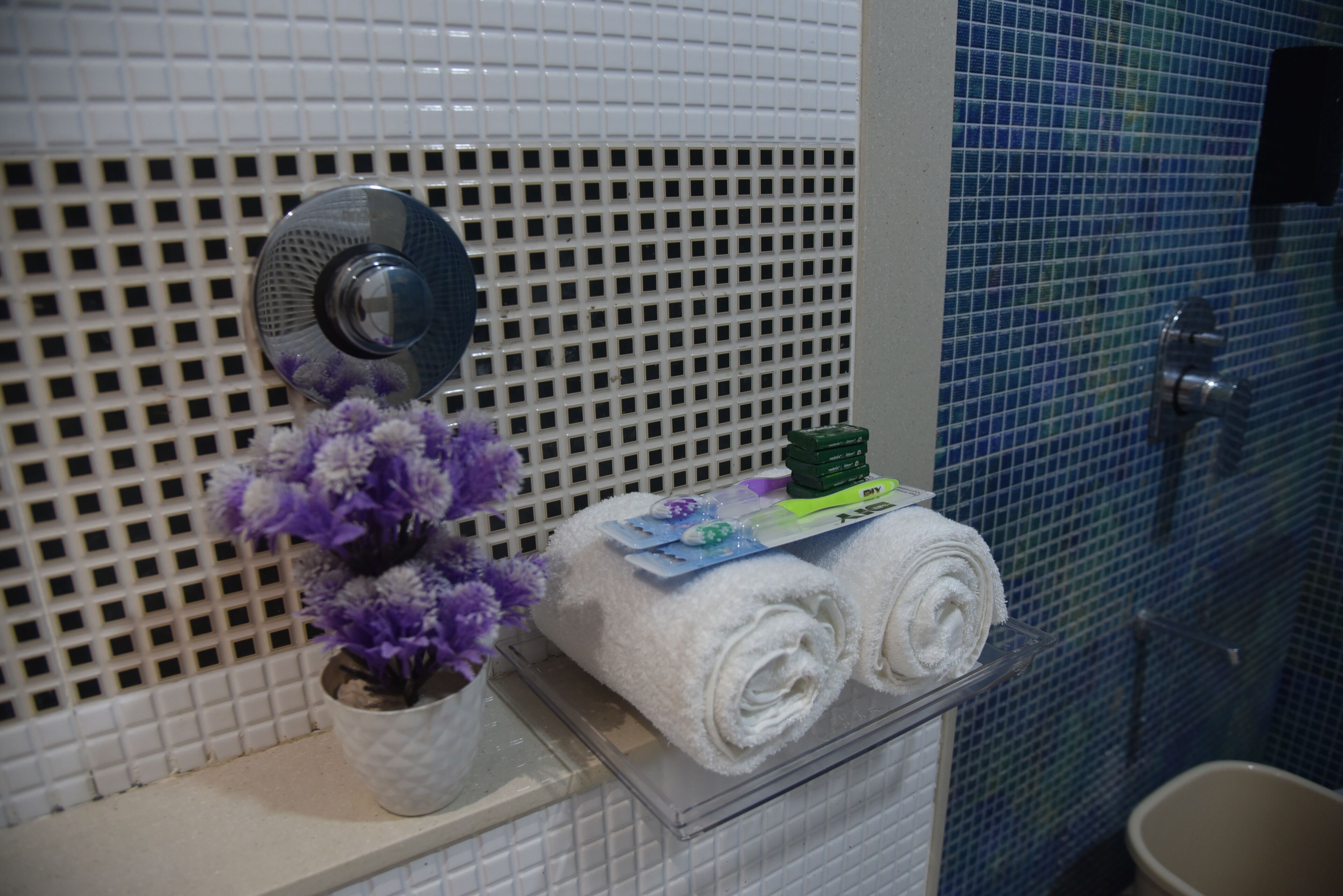 A bathroom with a white tiled wall, a shower area with blue mosaic tiles, a white sink with a mirror, and a towel rack with rolled towels and a potted plant.