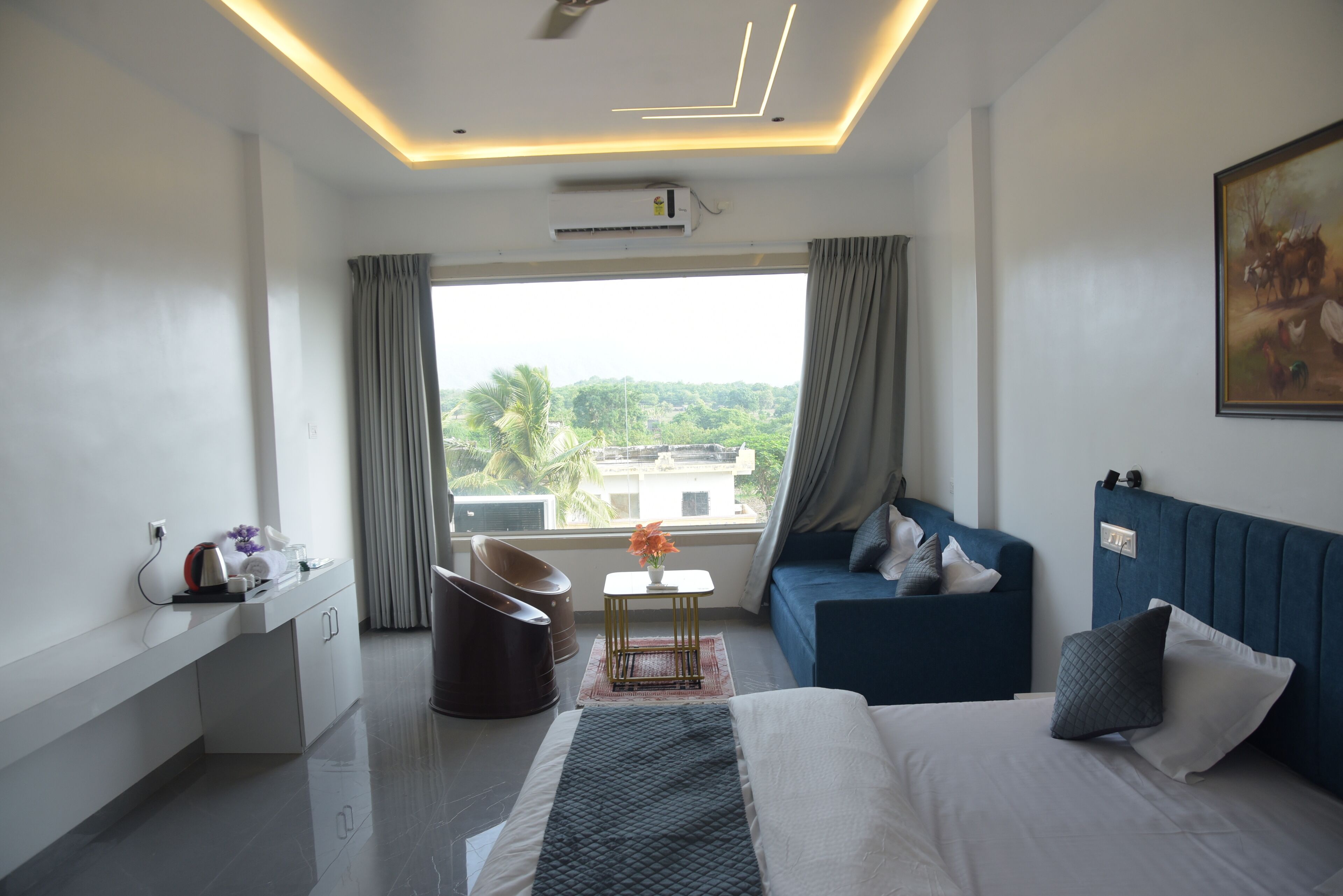 Ein modernes Hotelzimmer mit einem Bett, einem blauen Sofa, einem Schreibtisch mit Stuhl, einem kleinen Tisch mit einer Vase und einem großen Fenster mit Vorhängen.