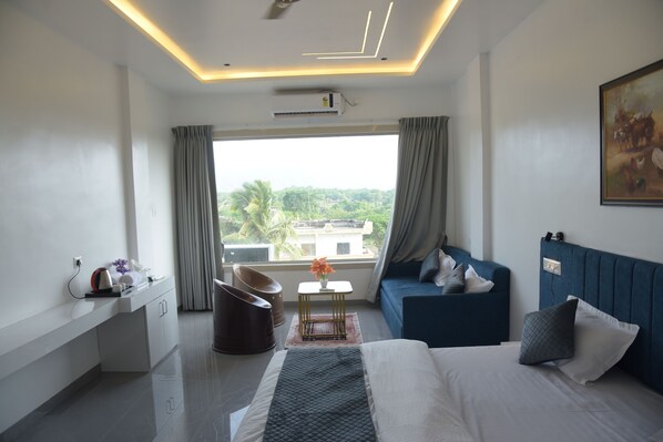 A modern hotel room with a bed, a blue sofa, a desk with a chair, a small table with a vase, and a large window with curtains.
