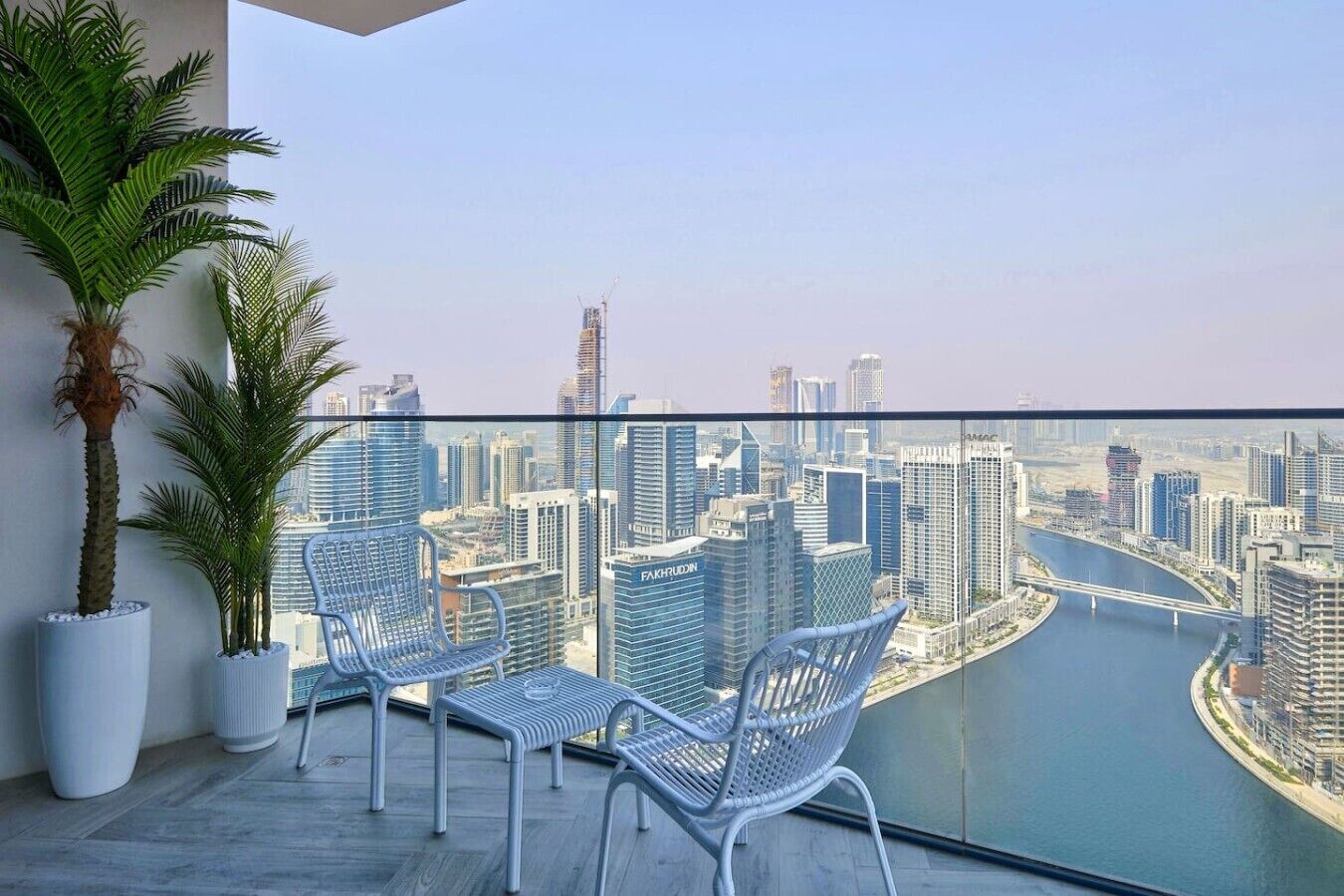 A balcony with two white wicker chairs, a potted palm tree, and a view of a cityscape with a river.