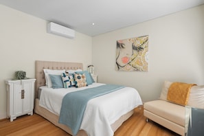 A bedroom with a bed, bedside tables, a nightstand, a chair, a wall-mounted air conditioner, and a painting. - Bella Rye by Ready Set Stay (Rye)