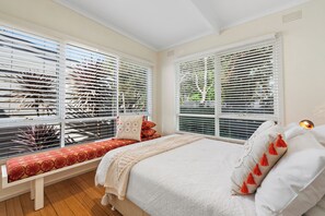Ein Schlafzimmer mit einem großen Bett, einer Bank mit roten Kissen und Blick durch weiße Jalousien.