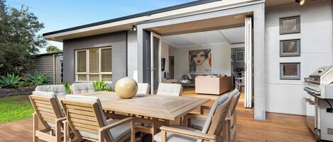 An outdoor patio with a wooden table and chairs, a barbecue grill, and a view into the house.