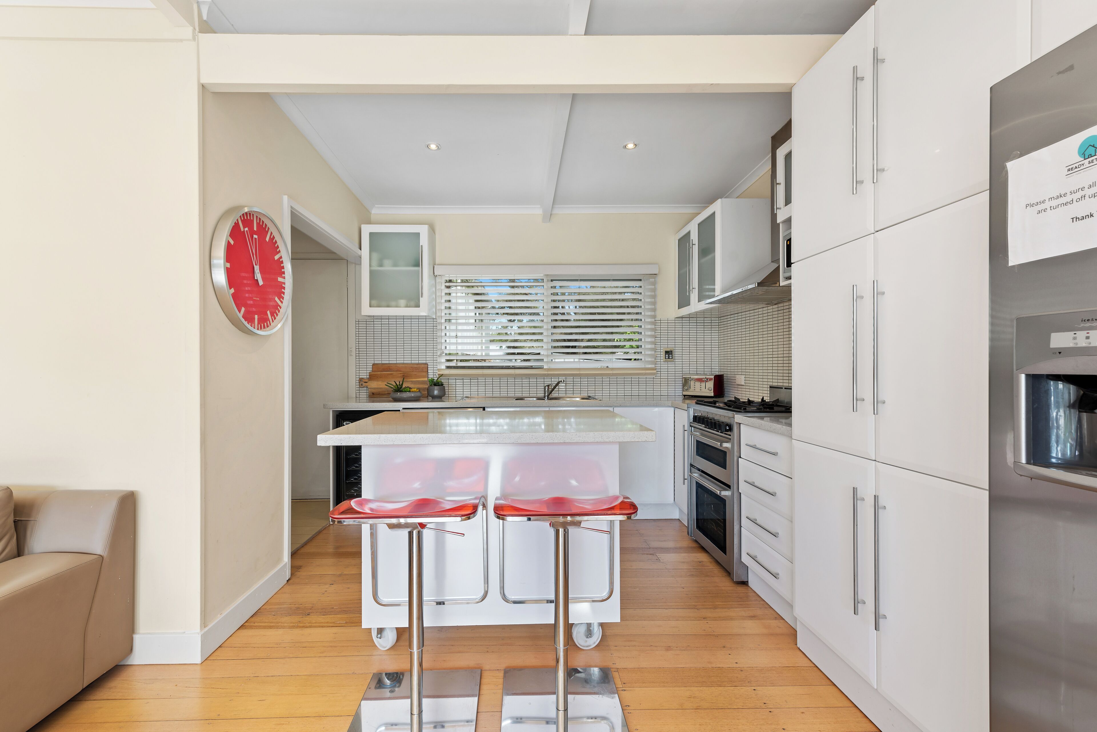 A modern kitchen with a central island, bar stools, white cabinetry, and a red clock on the wall.