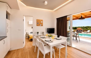 Eine moderne Küche mit weißen Schränken, ein Essbereich mit weißen Stühlen, ein Fernseher und Blick auf einen Pool.