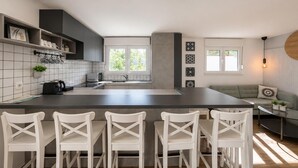 Een moderne keuken met een centraal eiland, barkrukken, een eethoek met een bank en uitzicht op groen.