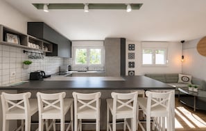 A modern kitchen with a central island, bar stools, a dining area with a sofa, and a view of greenery outside. - Amazing home in Zerava (Zerava)