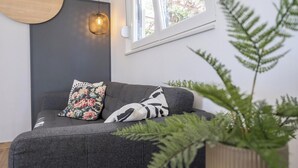 A modern living room with a grey sofa, two patterned cushions, a hanging light fixture, a window with a view, and a large green plant. - Stunning home in Zerava (Zerava)