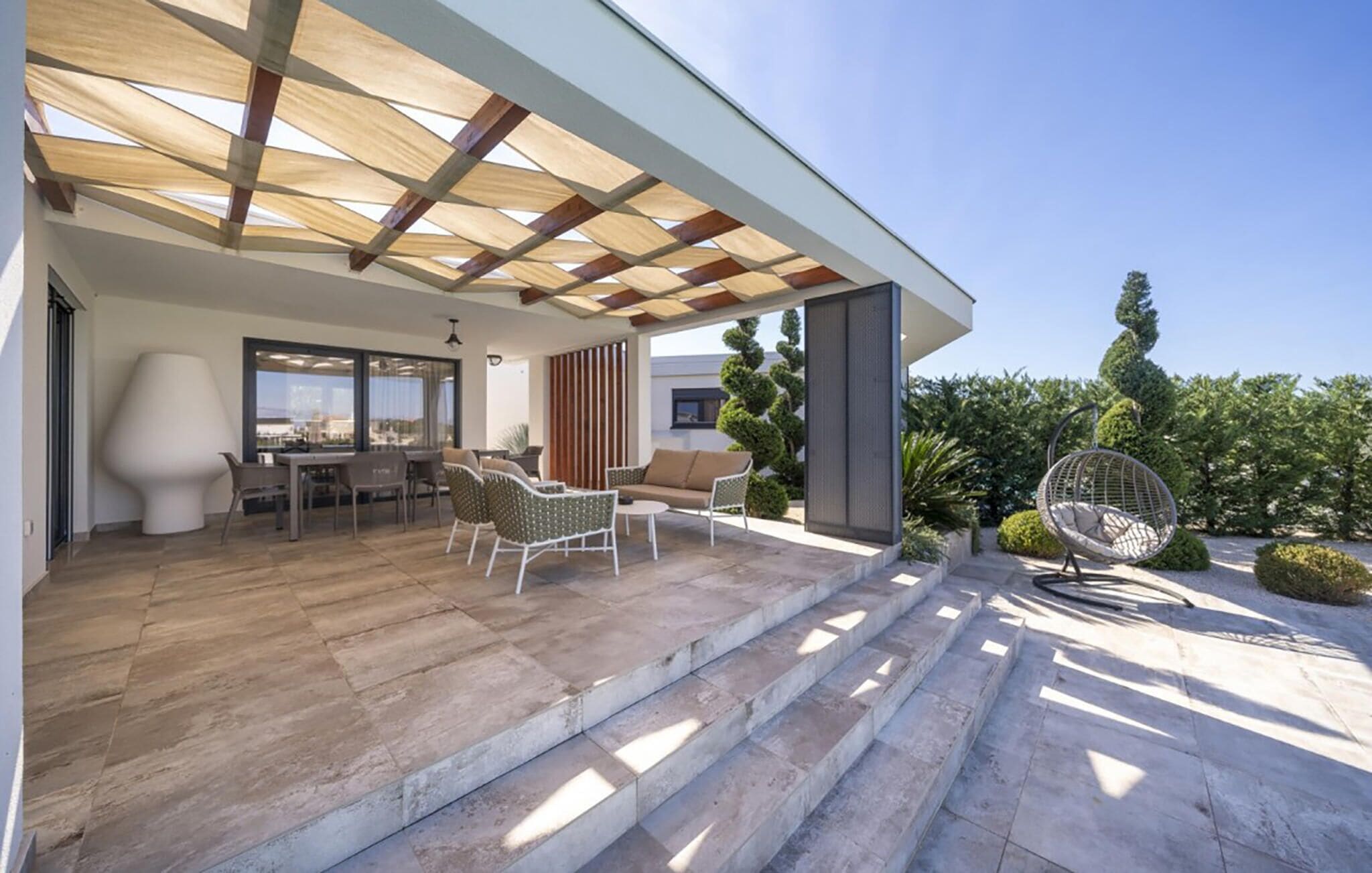 A modern outdoor patio with a pergola, seating area, and steps leading to a covered entrance.