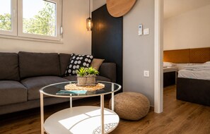 Ein modernes Wohnzimmer mit einer Couch, einem runden Glastisch, einem runden Hocker, einer Wandleuchte und einem Bett mit einem Holzkopfstück.