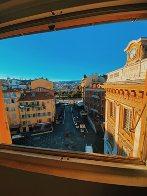 Blick aus einem Fenster auf eine Stadtstraße mit Gebäuden, einem Glockenturm und blauem Himmel.