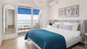 A bedroom with a large bed, a view of the sea through a sliding door, a ceiling light, and a painting on the wall. - Varandas do Mar - Estoril Atlantic Views (Cascais)