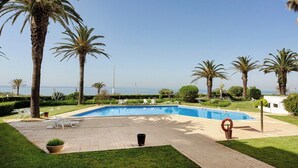 A pool area with a clear view of the sea, surrounded by palm trees and lounge chairs. - Varandas do Mar - Estoril Atlantic Views (Cascais)