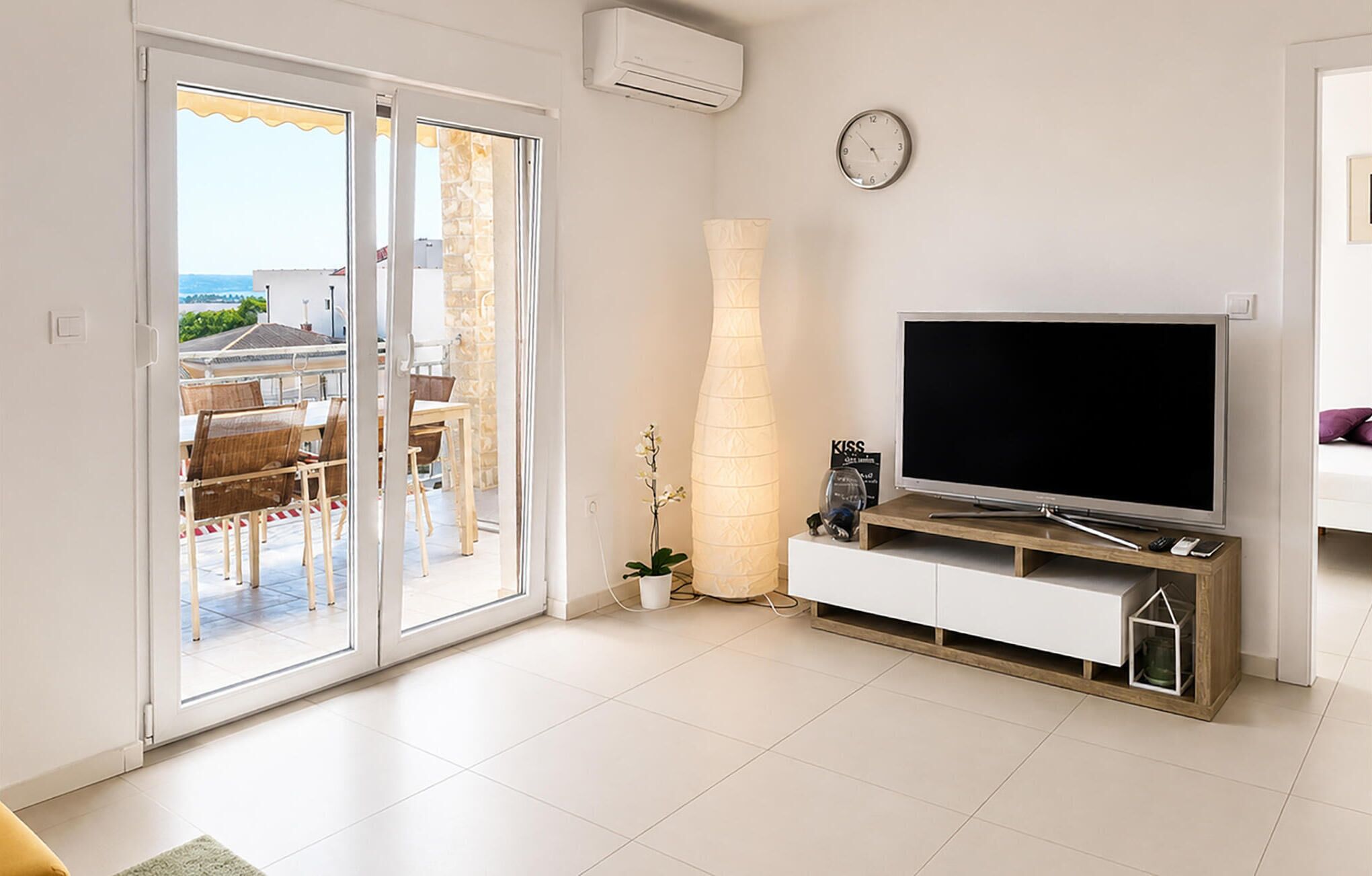 A living room with a TV, a clock, and a view of the outdoors through a sliding door.