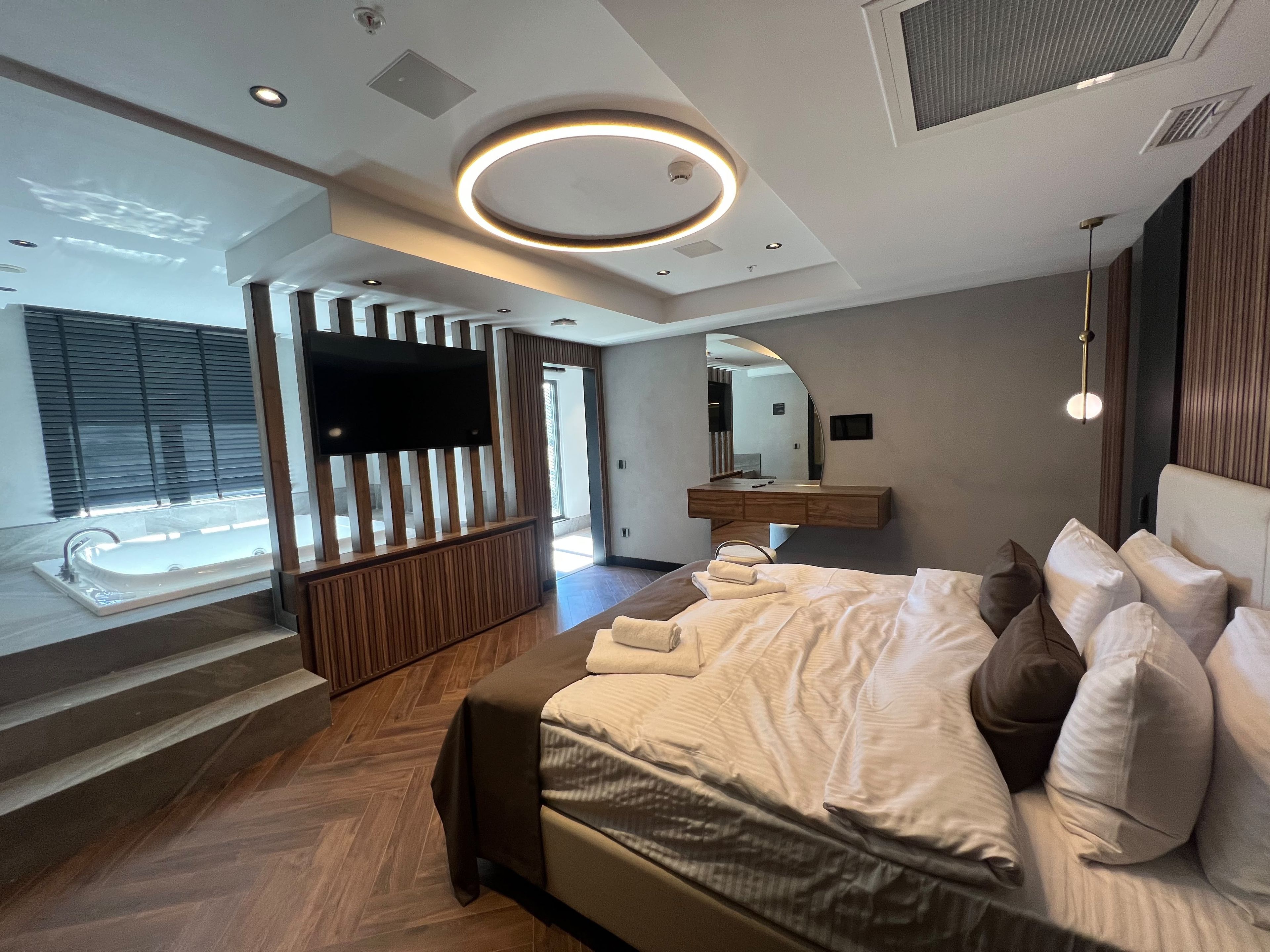 A modern bedroom with a large bed, a TV mounted on a wooden panel, and a bathroom visible through an open door.