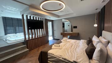 A modern bedroom with a large bed, a TV mounted on a wooden panel, and a bathroom visible through an open door.