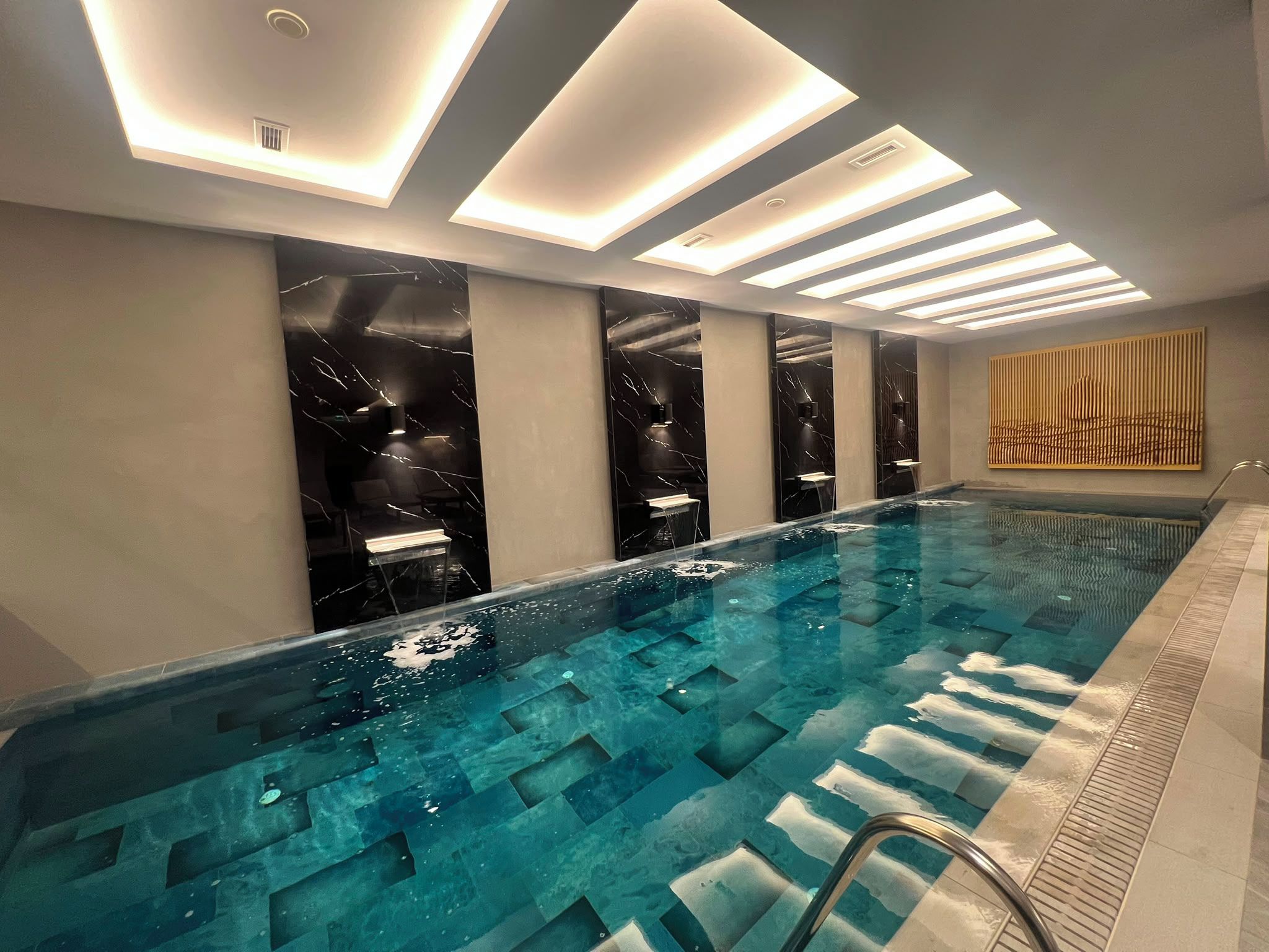 A modern indoor swimming pool with a clear water surface, a metal ladder, and a series of recessed lighting panels on the ceiling.